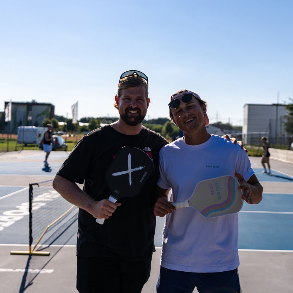 Die beiden Gründer von PIBA Sports halten Pickleball-Paddles von PIBA Sports und lachen in die Kamera, während sie auf den Pickleball-Plätzen von PIBA Neumarkt stehen – Leidenschaft für Pickleball und hochwertige Ausrüstung aus Deutschland