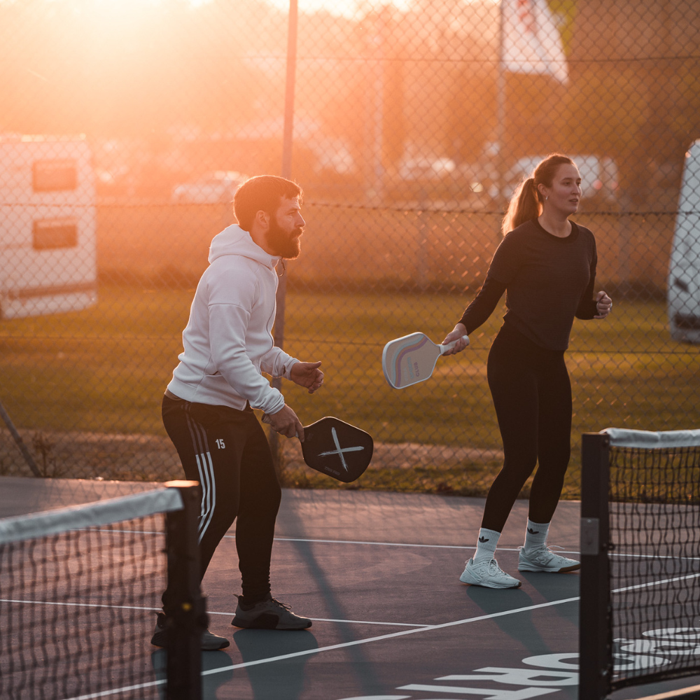 Zwei fokussierte Pickleball-Spieler – eine Frau und ein Mann – spielen Pickleball-Doppel bei Sonnenuntergang auf deutschen Pickleball-Plätzen, halten PIBA Sports Pickleball-Paddles und nutzen Pickleball Pro Netze.