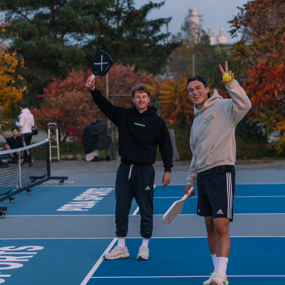 Zwei Pickleball-Spieler halten PIBA Sports Pickleball-Schläger in der Hand, lachen in die Kamera und stehen auf einem Pickleball-Platz in Deutschland – Freude am Pickleball mit hochwertiger Ausrüstung von PIBA Sports.